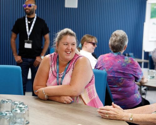 Sight Loss Council member Leanne is sat at a round table. She is smiling. Volunteers can be seen in the background, alongside TPT staff member Yahya.