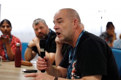 Merseyside SLC member Mike Spriggs holding a microphone talking to other SLC members at the 2025 SLC Volunteer Conference. He is sat at a roundtable with delegates, with other volunteers sat in the background listening.