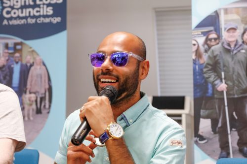 Sight Loss Council Engagement Manager Yahya speaks into the microphone at the SLC Conference 2025. SLC banners are in the background.