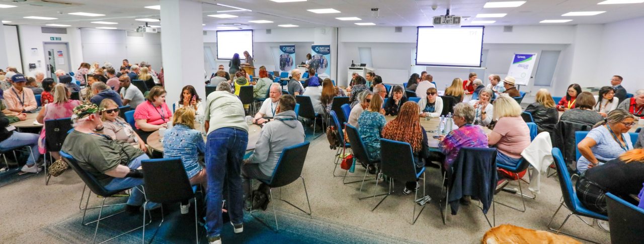 Sight Loss Council conference delegates (SLC volunteers) and sighted guides sat around round tables int he main conference hall. SLC banners are in the background, flip chart two larges screens to show the presentations. A golden coloured guide dog is laid on the floor.