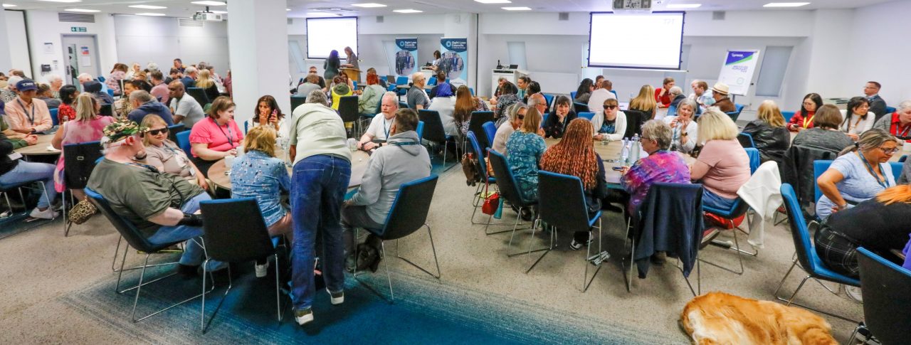 Sight Loss Council conference delegates (SLC volunteers) and sighted guides sat around round tables int he main conference hall. SLC banners are in the background, flip chart two larges screens to show the presentations. A golden coloured guide dog is laid on the floor.
