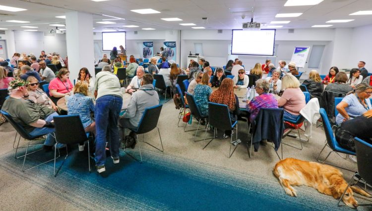 Sight Loss Council conference delegates (SLC volunteers) and sighted guides sat around round tables int he main conference hall. SLC banners are in the background, flip chart two larges screens to show the presentations. A golden coloured guide dog is laid on the floor.