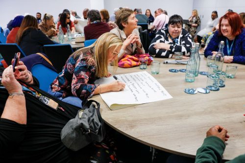 Volunteers engage in round-table discussions about making health accessible. Three of the tables can be seen in shot. A sighted guide is making notes of key points on flip chart paper.