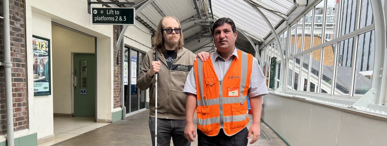 Attendee Gus is being guided by Noor at Sutton railway station. Gus is on the left, wearing black glasses and holding a long cane. He is wearing black shoes, black, denim jeans, and a brown zip-up hoodie. He has shoulder-length brown hair and a beard. Noor is on the right and is guiding Gus through the station. Noor is wearing his uniform, which is black shoes, smart black trousers, a light-coloured smart shirt, and an orange high-visibility jacket with his name badge on it. He has short, dark hair. They are both facing the camera.