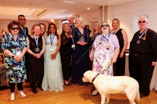 Birmingham and Black Country Sight Loss Council members group shot at the Rodney Powell Awards for Volunteering. Senior Engagement Manager Louise Connop is in centre holding the Team of the Year award. Everyone is smiling a the camera. A guide dog is also in the photo.
