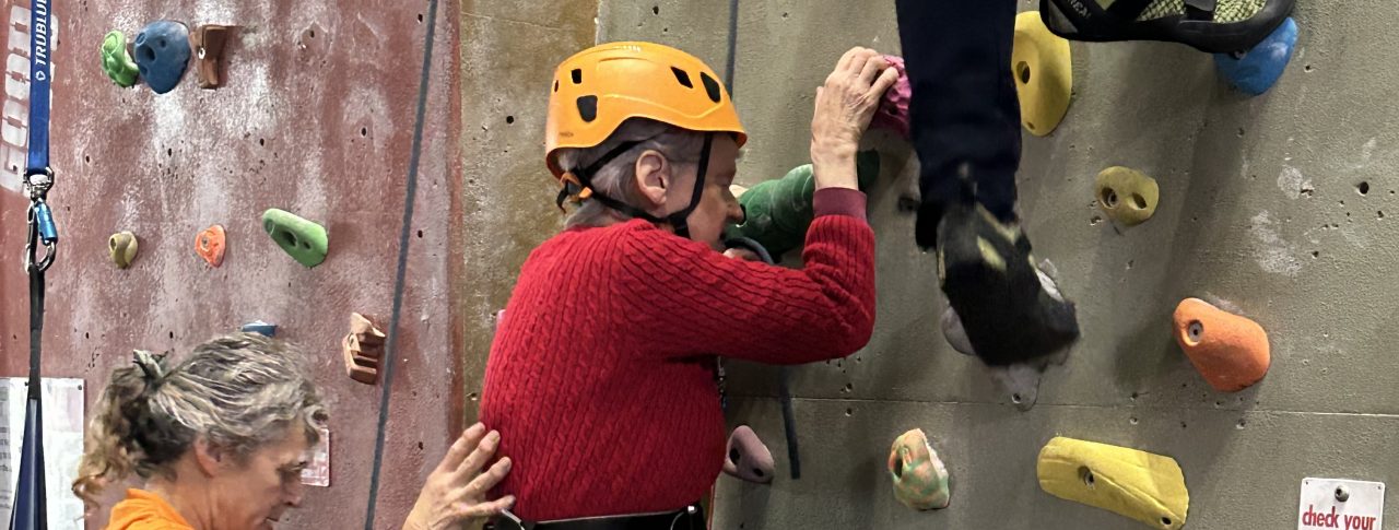 Two people wearing colourful helmets climbing the climbing wall at a previous 'Let's Get Active' event. A third lady, wearing a yellow sweat-shirt, stands at the bottom to support the climbers.