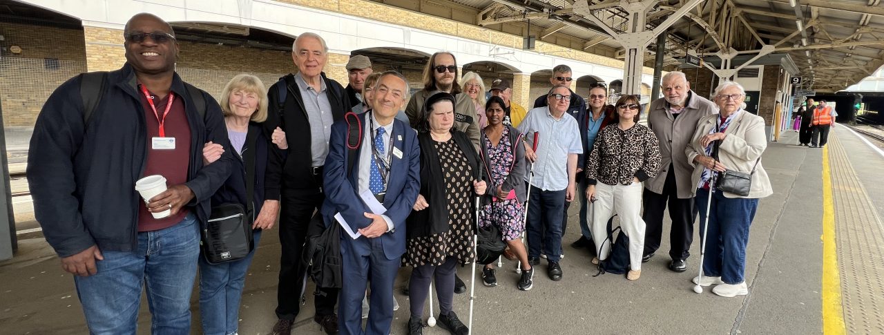 A group shot of 17 people of varying ages, genders, and ethnicities standing together at Wimbledon station. Five people are holding their long canes. Some people are carrying rucksacks, handbags, and coffee cups. Antony Merlyn stands at the front of the group in the middle, smiling at the camera. He is smartly dressed in a navy suit and tie, is holding a notebook, and carrying a rucksack.