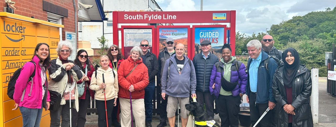 A group of seven people of mixed ages, genders and ethnicities. Some are holding white canes and there is one person with a guide dog. The group is standing in front of a red sign that reads, 'South Fylde Line' at a train station. There is an Amazon locker to the left and trees in the background.