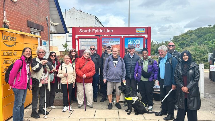 A group of seven people of mixed ages, genders and ethnicities. Some are holding white canes and there is one person with a guide dog. The group is standing in front of a red sign that reads, 'South Fylde Line' at a train station. There is an Amazon locker to the left and trees in the background.