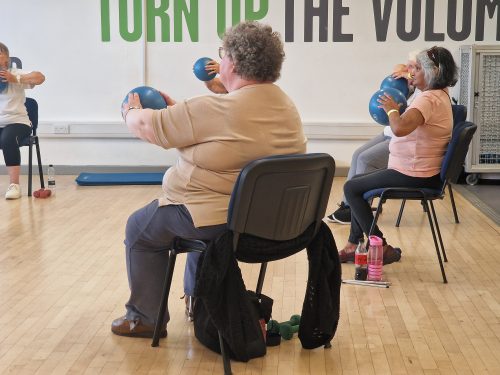 Five delegates sitting on chairs inside a sports hall, with the three at the front most visible. They are holding blue balls, which they are using to exercise. They are dressed in comfortable clothing, and there are water bottles, and dumbbells on the floor by their chairs. It says 'Turn up the volume' in big green and black capital letters on the back wall.