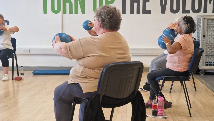 Five delegates sitting on chairs inside a sports hall, with the three at the front most visible. They are holding blue balls, which they are using to exercise. They are dressed in comfortable clothing, and there are water bottles, and dumbbells on the floor by their chairs. It says 'Turn up the volume' in big green and black capital letters on the back wall.