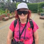 A headshot of Helen Lawson. Helen is standing up wearing a pink t-shirt, a grey hat, a visor, a lanyard around her neck and a backpack.