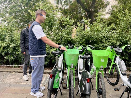 Hal Stevenson, Director of Policy (UK and Ireland), approaching a bay of parked e-bikes, whilst in sim-specs, during the walk. He is wearing grey trousers and a white t-shirt and grey gilet. 