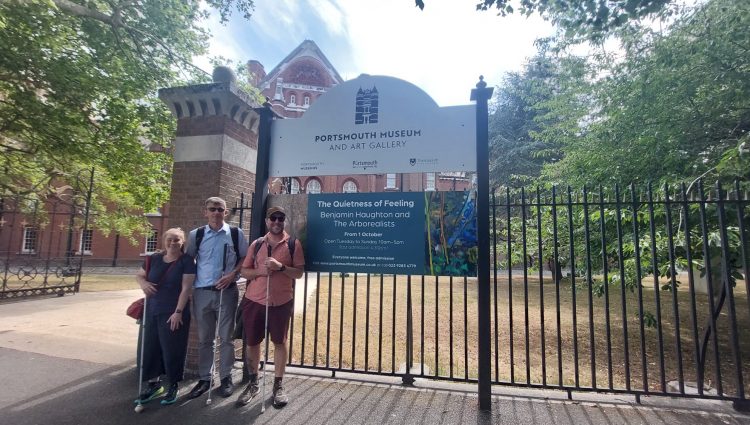 Three people, a woman and two men, are standing slightly to the left of a sign in front of a large building. The people are all holding white walking sticks, and the two men in the centre and to the right are wearing visors. The sign the people are standing next to says ‘Portsmouth Museum and Art Gallery” while an additional sign below is promoting an event for, ‘The Quietness of Feeling’ on the 1st of October.