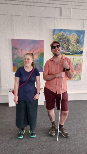 Two attendees, a woman and a man, are standing in front of some artwork that is fixed to a white wall in the background. Both are smiling at the camera.
