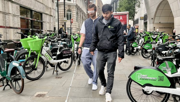 Two men in the centre. The man on the left is wearing a navy gilet and is being guided by the other man who is wearing a black jacket down a narrow street due to a plethora of poorly parked e-bikes. The e-bikes are scattered either side of the two men as the man on the left uses a walking stick and special visors to help navigate the street.