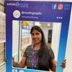 Shalini O'Kane headshot: Shalini stands inside an optician's store, holding a large promotional frame in blue and purple colours. The frame features branding for 'Rawlings Opticians & Hearing Care' and includes social media handles, hashtags, and cartoon icons of an ear and an eye.