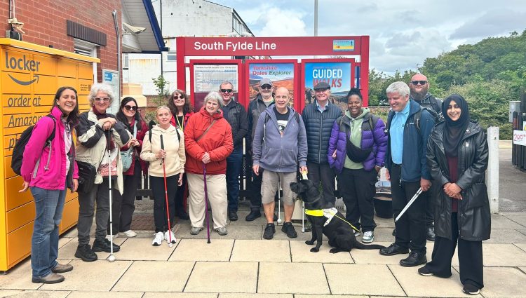 A group of seven people of mixed ages, genders and ethnicities. Some are holding white canes and there is one person with a guide dog. The group is standing in front of a red sign that reads, 'South Fylde Line' at a train station. There is an Amazon locker to the left and trees in the background.