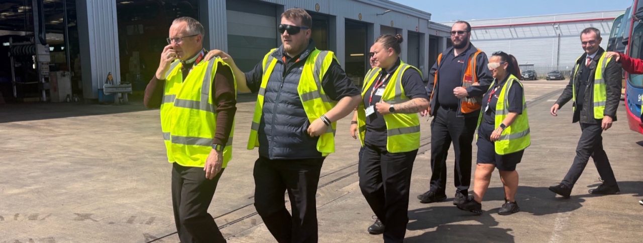 A group of Go North East staff are walking together in a line outside a bus depot. They are all wearing high-visibility vests and are participating in a visual impairment awareness exercise. The person at the front is leading with their hand raised, while the others are following with their hands on the shoulder of the person in front. Some participants are wearing simulation glasses to replicate various sight conditions. Large garage bays and buses are visible in the background, under a clear blue sky. The mood is focused and collaborative.
