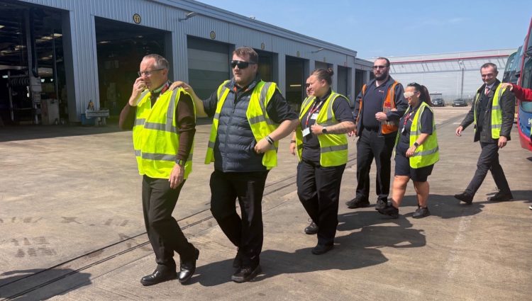 A group of Go North East staff are walking together in a line outside a bus depot. They are all wearing high-visibility vests and are participating in a visual impairment awareness exercise. The person at the front is leading with their hand raised, while the others are following with their hands on the shoulder of the person in front. Some participants are wearing simulation glasses to replicate various sight conditions. Large garage bays and buses are visible in the background, under a clear blue sky. The mood is focused and collaborative.