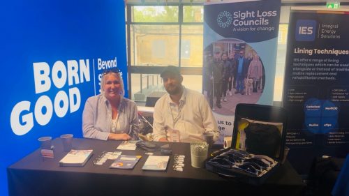 Volunteer Emma and engagement manager Jack Moffat are seated behind a Sight Loss Councils exhibition table at an indoor event. They are smiling at the camera with informational leaflets, lanyards, and simulation glasses neatly displayed on the table in front of them. Behind them are three tall banners: one with bold text reading “BORN GOOD | Beyond” on a bright blue background, a second featuring the Sight Loss Councils logo and a group photo, and a third from Integral Energy Solutions (IES) promoting lining techniques. A large window in the background lets in natural light, creating a welcoming and vibrant atmosphere at the event.