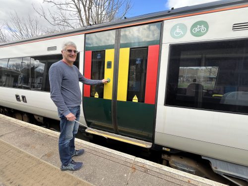 SLC member Clinton is standing next to a trains doors and is pushing the open doors button. Clinton is looking at the camera smiling, while holding a white-cane.