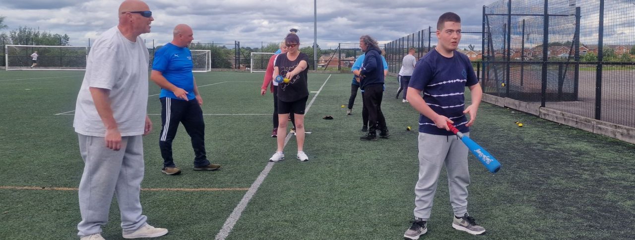A group of six attendees at a previous Let's Get Active event learning and practising baseball on an outdoor pitch. To the right of the image, a young male attendee holding a bat stands slightly in front of the rest of the players.