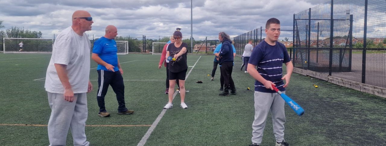 A group of six attendees at a previous Let's Get Active event learning and practising baseball on an outdoor pitch. To the right of the image, a young male attendee holding a bat stands slightly in front of the rest of the players.