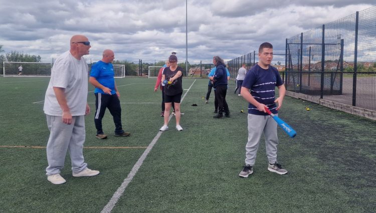 A group of six attendees at a previous Let's Get Active event learning and practising baseball on an outdoor pitch. To the right of the image, a young male attendee holding a bat stands slightly in front of the rest of the players.