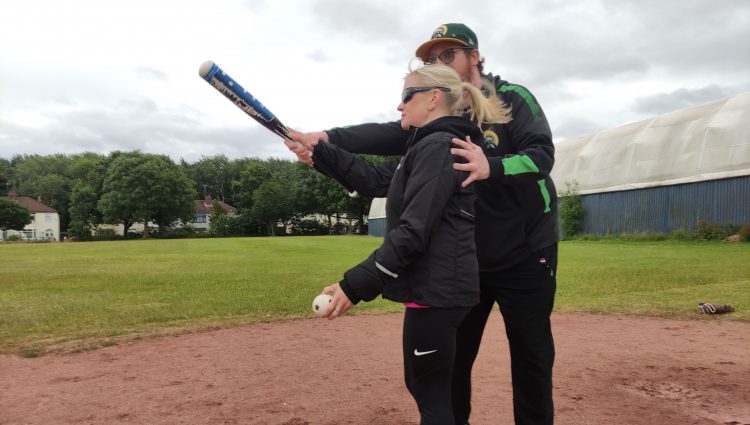 Kelly Barton, Engagement Manager for the North West during a VI Baseball session. She is standing on the field at the home base. She is with an instructor who is guiding her where to position the bat. Kelly is holding the baseball in her other hand.