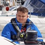 Photo of Lloyd Sakr, Lancashire SLC member. Lloyd is sitting behind ropes on a sail boat, wearing a blue jacket.