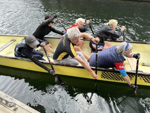 SLC volunteer Mike Wright and his crew having just completed a Dragon Boat training session at Queens Dock Liverpool. Mike travels to his training sessions and races by train. There is a group of six people in a yellow and black dragon boat, three at either side, rowing with oars. Mike is on the nearside row in the middle. 