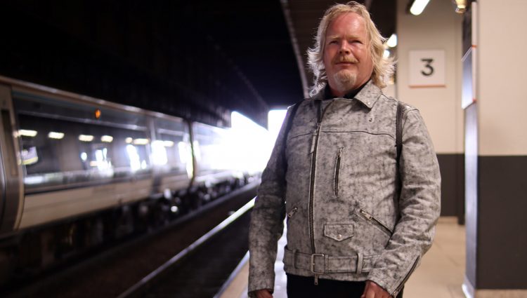 Portrait image of Steve Keith, Birmingham and Black Country SLC member, standing on a train platform with his guide dog. A train is on the opposite side of the tracks.