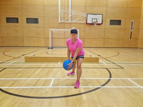 Engagement Manager Kelly is pictured playing goalball. She is dressed in pink sportswear and is wearing a blindfold, standing poised with a blue foam ball in her hand.