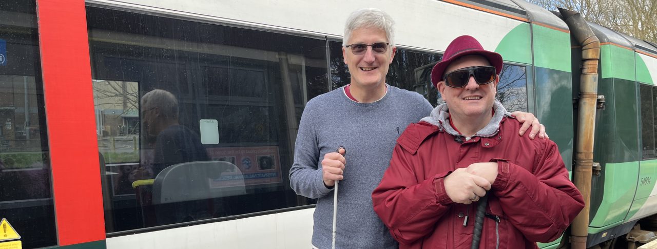 Two SLC members, Clinton on the left and Paul on the right, are standing together holding canes and smiling at the camera. They are standing in front of a green and white train.