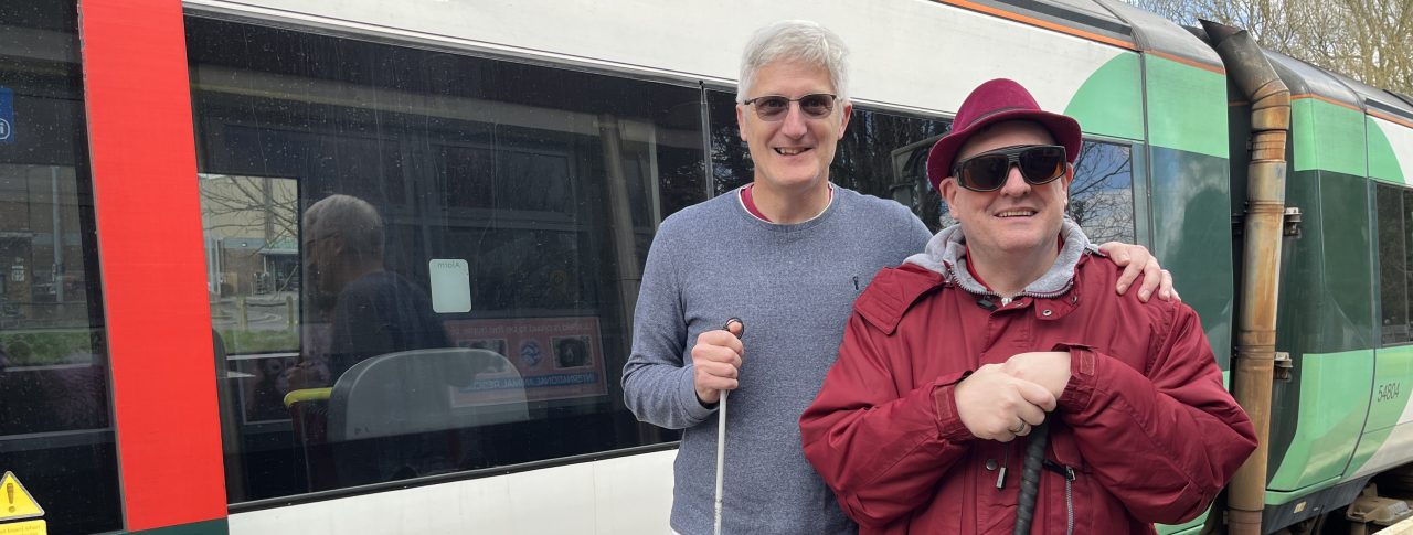 Two SLC members, Clinton on the left and Paul on the right, are standing together holding canes and smiling at the camera. They are standing in front of a green and white train.