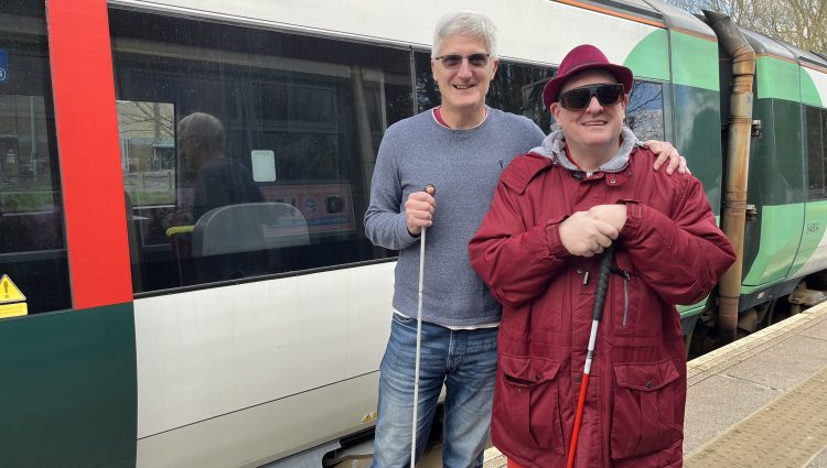 Two SLC members, Clinton on the left and Paul on the right, are standing together holding canes and smiling at the camera. They are standing in front of a green and white train.